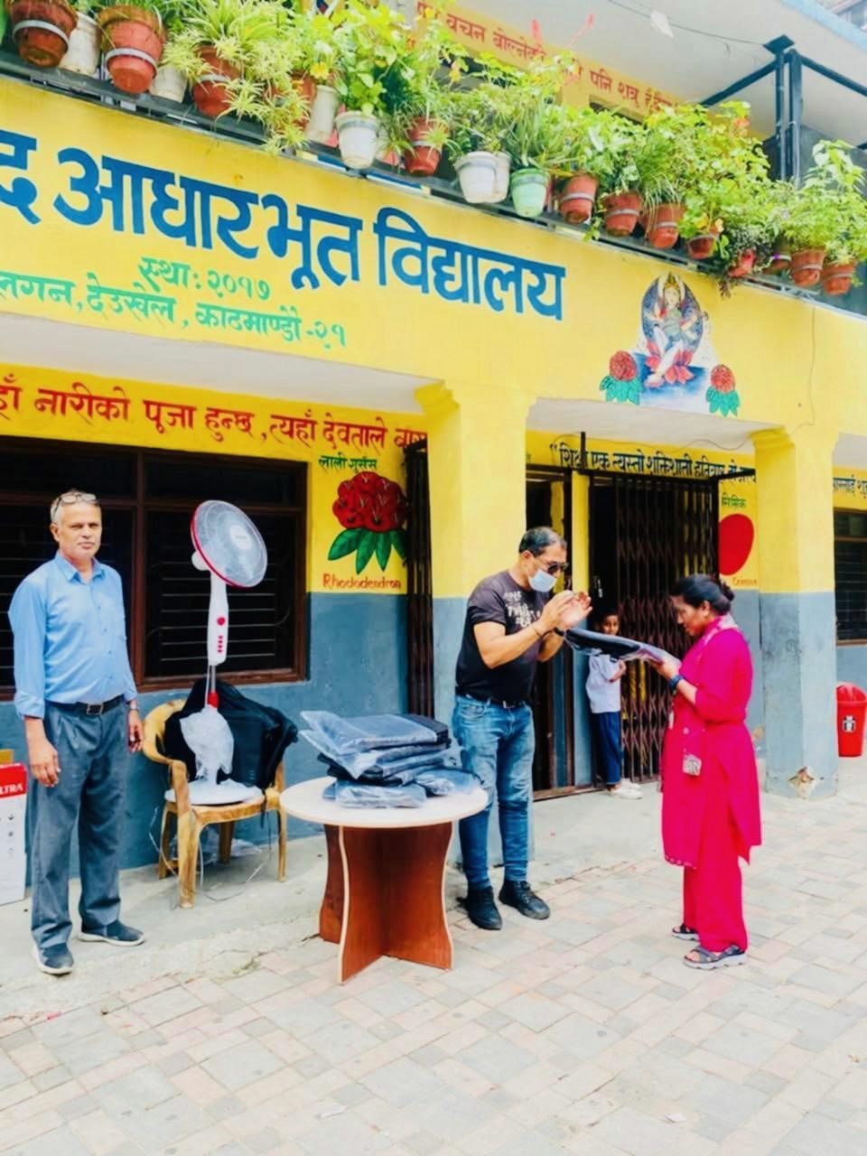 Three people presenting a classroom fan outside a yellow school building